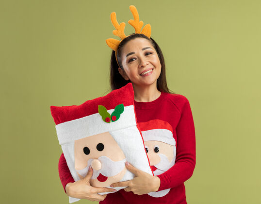 角穿着红色圣诞毛衣的快乐年轻女子 戴着滑稽的鹿角边 抱着圣诞枕头 面带微笑圣诞有趣的鹿