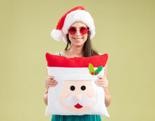 眼镜戴着太阳眼镜 戴着圣诞帽 抱着圣诞枕头的年轻高加索女孩太阳复制举行