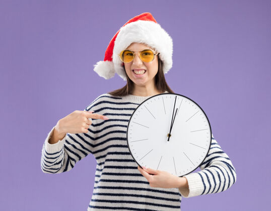拿着戴着太阳眼镜 拿着圣诞帽 指着时钟的年轻白种人女孩新的年新年