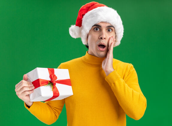 圣诞节一个穿着黄色高领毛衣 戴着圣诞老人帽 打着滑稽领带的年轻人拿着一份礼物 站在绿色的墙边惊讶地看着站着领带惊讶