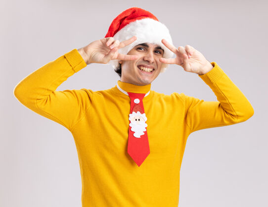 靠近滑稽的年轻人穿着黄色高领毛衣 戴着圣诞老人帽 打着滑稽的领带 眼睛附近有个v形标志 站在白色的墙上开心地微笑着圣诞节圣诞老人站着