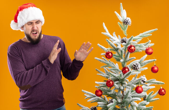 男人一个穿着紫色毛衣 戴着圣诞帽的年轻人站在橙色的墙上 厌恶地看着圣诞树做着防御的手势表情圣诞节厌恶