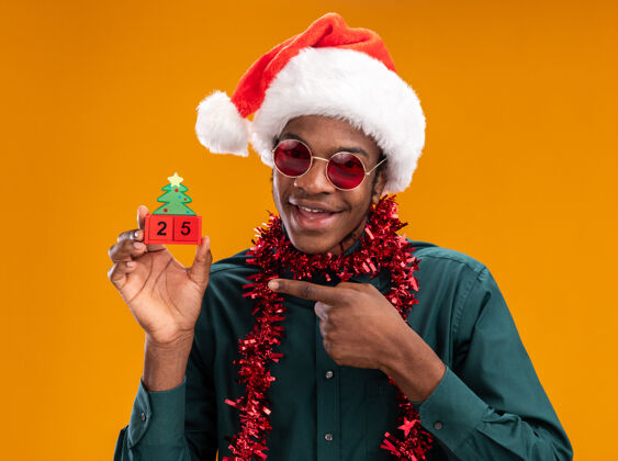 手指快乐的非洲裔美国人戴着圣诞帽 戴着花环 戴着太阳镜 手里拿着日期为25的玩具方块 用食指指着站在橘色墙上的玩具方块日期戴着帽子