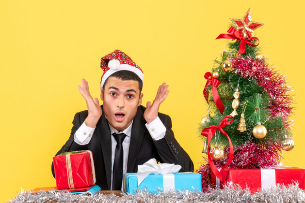桌子前视图：穿着西装 戴着圣诞帽的男人坐在桌子旁 圣诞树和礼物穿西装的人帽子商人