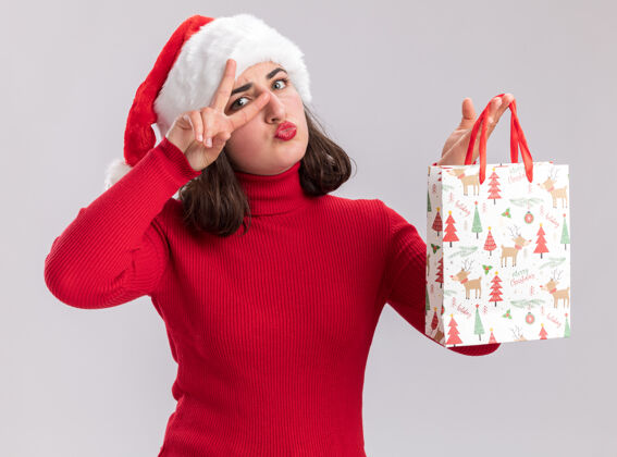 圣诞老人快乐的年轻女孩穿着红色毛衣 戴着圣诞帽 手里拿着彩色纸袋 里面放着圣诞礼物 眼睛旁边有个v形标志 站在白色的墙上Vsign展示帽子