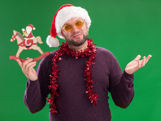 帽子高兴的中年男子戴着圣诞老人帽 脖子上戴着金箔花环 戴着眼镜 把圣诞老人抱在摇摇木马上手周围花环
