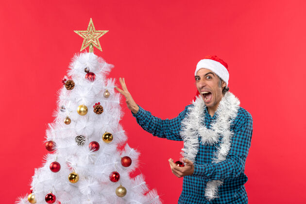 年轻人一个戴着圣诞老人帽子的紧张的年轻人 穿着一件蓝色的衬衫 在红色的圣诞树上装饰圣诞树圣诞老人圣诞老人圣诞树