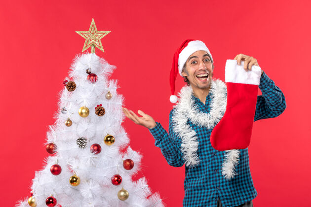男人兴奋的快乐的年轻人戴着圣诞老人的帽子 穿着蓝色的衬衫 手里拿着圣诞袜 靠近圣诞树兴奋圣诞老人圣诞老人