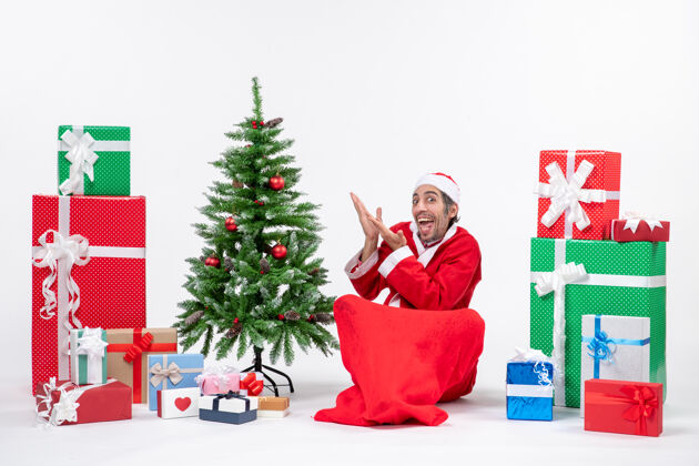 指年轻人打扮成圣诞老人 拿着礼物和装饰好的圣诞树坐在地上指着什么打扮礼物庆祝
