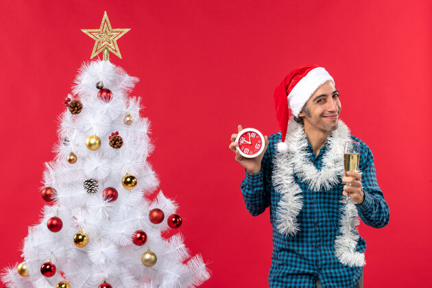 圣诞老人快乐骄傲的年轻人 戴着圣诞老人的帽子 举起一杯葡萄酒 拿着时钟站在圣诞树旁的红股票照片葡萄酒年轻人小伙子