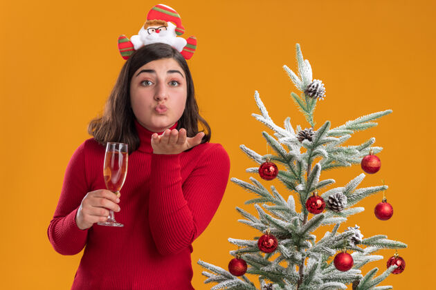 搞笑穿着圣诞毛衣的快乐小女孩戴着滑稽的头带 手里拿着一杯香槟 在橙色背景下的圣诞树旁吹吻毛衣年轻戴