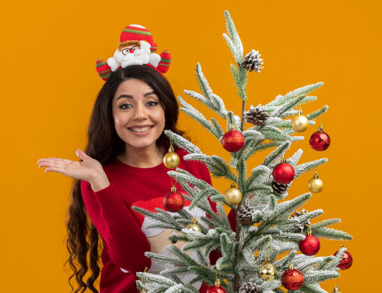 手微笑着的年轻漂亮女孩戴着圣诞老人的头带和毛衣站在装饰好的圣诞树后面看着相机 在橙色的背景上显示出孤立的空手圣诞老人快乐漂亮