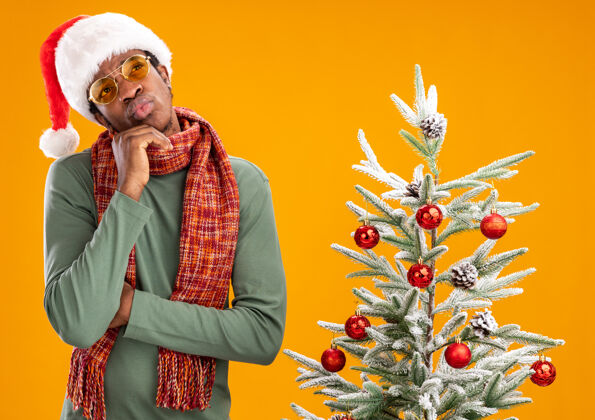 脖子戴着圣诞帽 脖子上围着围巾的非洲裔美国人站在橙色背景下的圣诞树旁 困惑地抬头望去看帽子圣诞