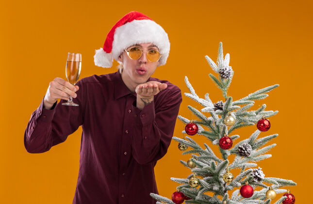 圣诞自信的金发女郎戴着圣诞帽 戴着眼镜 站在装饰好的圣诞树旁 手里拿着一杯香槟 看着摄像机 在橙色的背景下 孤零零地送上一个飞吻发送靠近帽子