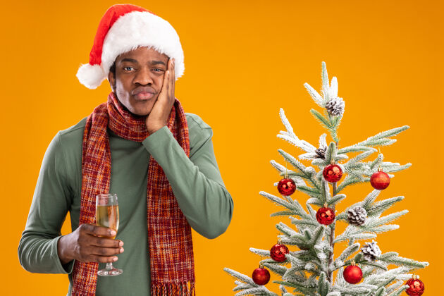 男人戴着圣诞帽 围着围巾的非洲裔美国男子拿着一杯香槟看着摄像机疲惫无聊地站在橙色背景下的圣诞树旁无聊站着相机