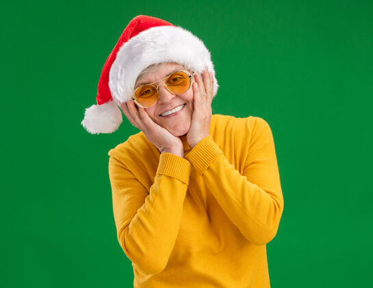 手戴着太阳眼镜 戴着圣诞帽 面带微笑的老妇人把手放在脸上 隔离在绿色背景上 留有复印空间眼镜圣诞老人老人