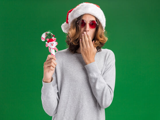 圣诞老人年轻人戴着圣诞老人帽 戴着红眼镜 拿着圣诞糖果手杖看着镜头 震惊地捂着嘴 手站在绿色的背景上帽子年轻手