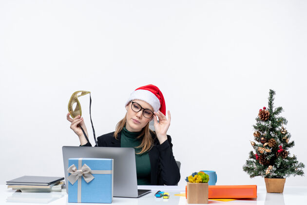 帽子戴着圣诞老人帽 眼镜和面具的年轻女子坐在桌子旁 手里拿着圣诞树和礼物笔记本笔记本商务