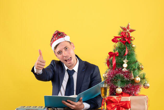 树商务人士在圣诞树旁的桌子旁做竖起大拇指的招牌 黄色的礼物黄色桌子制作