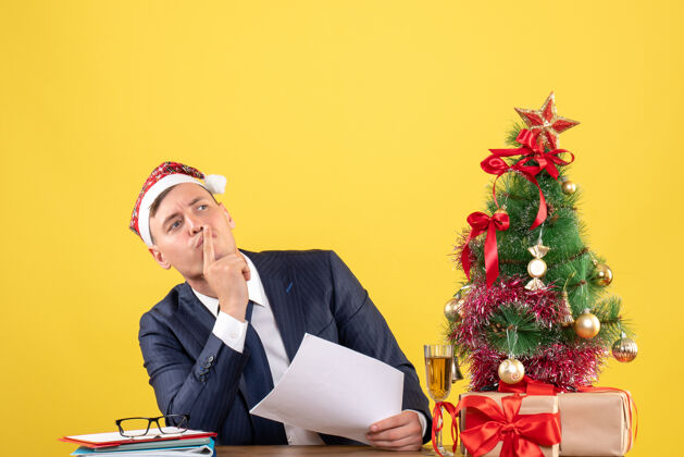 男人商务人士在圣诞树旁的桌子旁做嘘标志的正面图 黄色的礼物圣诞节新郎坐着