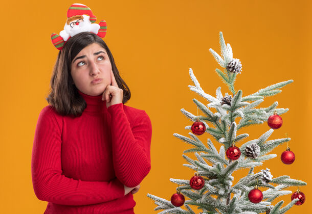 困惑穿着圣诞毛衣戴着滑稽头带的小女孩在橙色背景下的圣诞树旁困惑地抬头看有趣站着穿