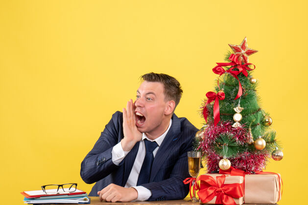 商人愤怒的男人坐在圣诞树旁的桌子前大声喊叫 黄色的墙上摆着礼物愤怒树生意