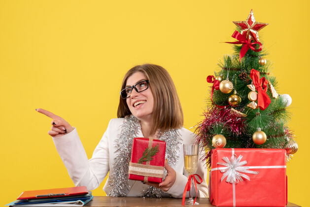 办公室正面图：女医生坐在桌子前 拿着礼物 黄色背景上的圣诞树和礼盒女医生情绪前面