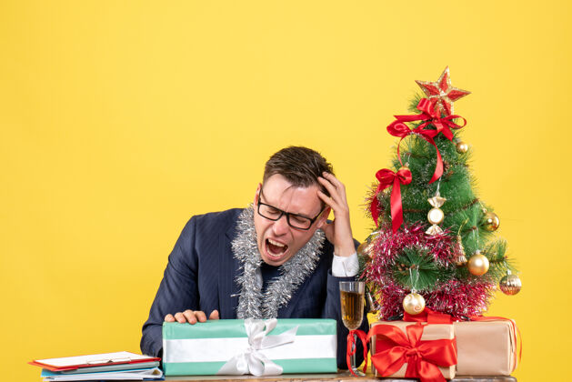 礼物愤怒的男人坐在圣诞树旁的桌子前 在黄色的墙上展示 并留有复印空间树前面工作