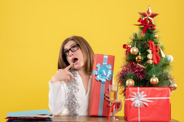 女正面图：女医生坐在桌子前 拿着礼物 黄色背景上的圣诞树和礼盒圣诞节树快乐