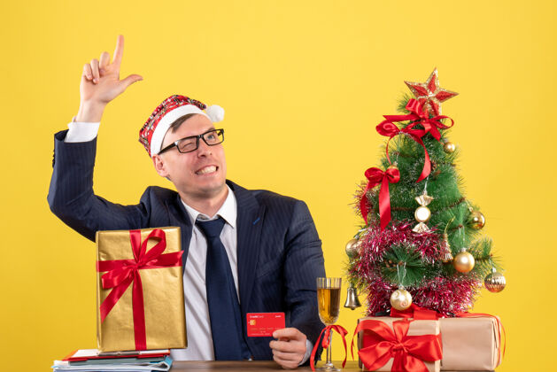 看商务人士戴着圣诞帽坐在圣诞树旁的桌子旁仰视着黄色的礼物帽子新郎圣诞节