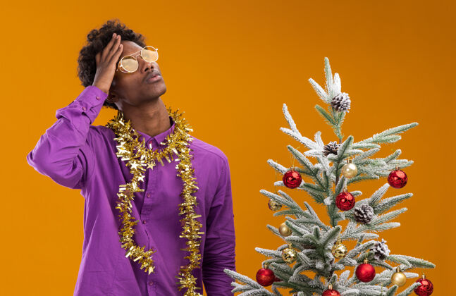 金属片困惑的年轻美国黑人男子戴着眼镜 脖子上戴着金箔花环 站在橙色背景的装饰圣诞树旁眼镜附近头