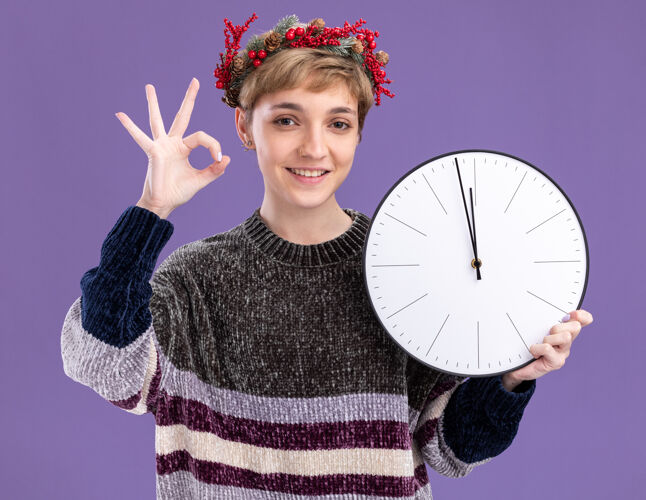 抱着微笑的年轻漂亮女孩戴着圣诞花环拿着时钟看着相机在紫色背景上做ok标志相机微笑年轻