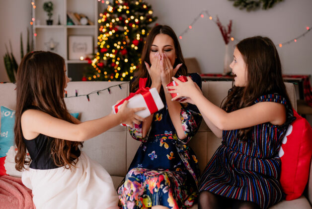 送印象深刻的两姐妹和年轻兴奋的妈妈在家过圣诞节的时候坐在客厅的沙发上姐妹们给妈妈送礼物包 她看着包手捂着嘴快乐保持嘴