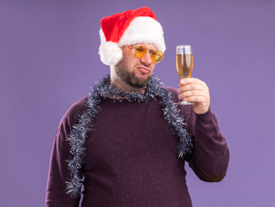 玻璃杯可疑的中年男子戴着圣诞老人的帽子 脖子上戴着金属丝的花环 戴着一副眼镜 手里拿着一杯孤立在紫色墙上的香槟帽子可疑花环