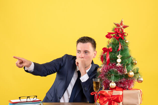 商人前视图好奇的人手指的东西坐在圣诞树和礼物旁边的黄色桌子上桌子西装好奇