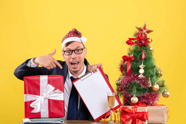 坐着前视图的商人指着剪贴板坐在桌旁圣诞树和黄色的礼物礼物圣诞节男