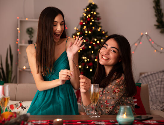 漂亮笑容可掬的年轻女孩拿着闪闪发光的灯 看着坐在桌边的相机 在家里享受圣诞节时光快乐时间火花