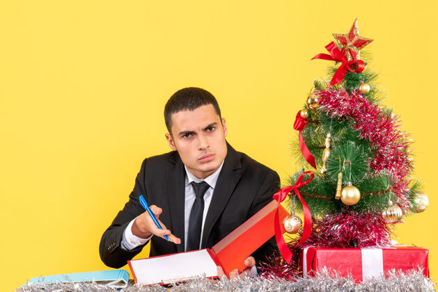 年轻人正面图：一个穿着西装的年轻人坐在桌子旁 展示圣诞树和礼物文件展示西装