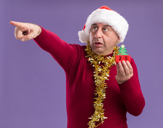 困惑中年男子戴着圣诞老人的帽子 脖子上戴着金属丝 手里拿着日期为25的玩具立方体 用食指指着站在紫色背景上困惑的东西圣诞老人日期帽子