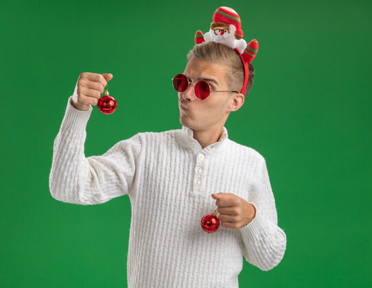 圣诞饰品可疑的年轻帅哥戴着圣诞老人的头带 戴着眼镜 手里拿着圣诞饰品 看着其中一个被隔离在绿墙上的复制品圣诞老人戴着圣诞老人