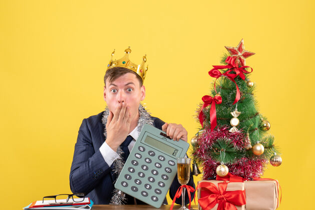 树前视图惊奇的人拿着计算器坐在圣诞树旁边的桌子上 黄色的墙上摆着礼物礼物男人键盘乐器