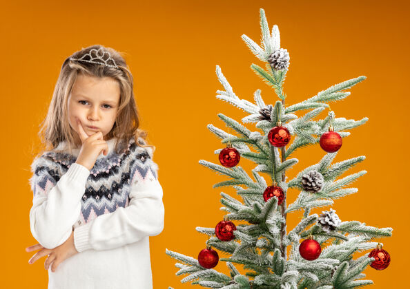 脖子想着站在圣诞树旁戴着头饰脖子上戴着花环的小女孩 手放在下巴上 孤立在橙色背景上女孩站立圣诞节