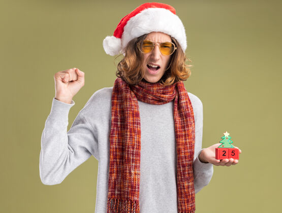 围巾年轻人戴着圣诞老人帽 戴着黄色眼镜 脖子上围着暖和的围巾 手里拿着和25岁约会的玩具立方体 紧握着拳头 站在绿色的背景上 快乐而兴奋脖子帽子圣诞节