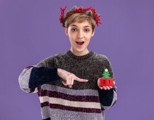 圣诞节令人印象深刻的年轻漂亮女孩戴着圣诞花环手持圣诞树玩具与日期指向它隔离在紫色墙上指向年轻印象深刻