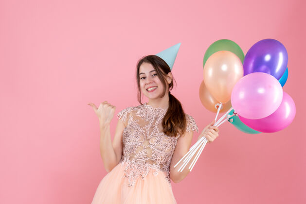 人快乐的派对女孩戴着派对帽拿着气球手指着粉色的东西漂亮举行帽子
