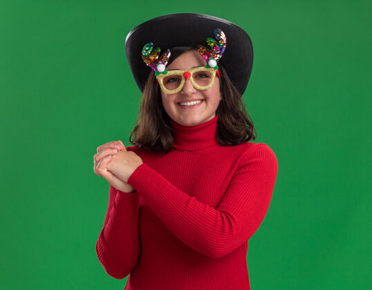 积极穿红毛衣的小女孩戴着滑稽的眼镜 戴着黑色的帽子 手拉着手 快乐而积极的微笑 欢快地站在绿色的墙上毛衣穿着圣诞节