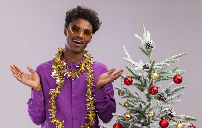 站着快乐的年轻黑人男子戴着眼镜 脖子上戴着金箔花环 站在装饰过的圣诞树旁 双手空空地孤零零地站在白色的墙上年轻附近空