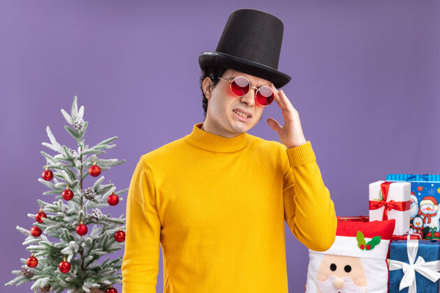 触摸一个穿着黄色高领毛衣戴着黑帽子戴着眼镜的年轻人 看起来很不舒服 他站在一棵圣诞树旁边摸着头 头痛 紫色的墙上挂着礼物站立帽子不舒服