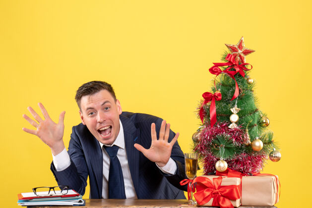 桌子正面图是快乐的男人张开双手坐在圣诞树旁的桌子旁 黄色的礼物礼物树西装