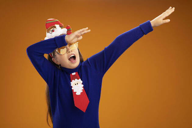 女孩小女孩穿着蓝色高领毛衣 打着红色领带 头上戴着有趣的圣诞戒指 开心地打着手势 双手站在橙色的背景上手势头高领毛衣